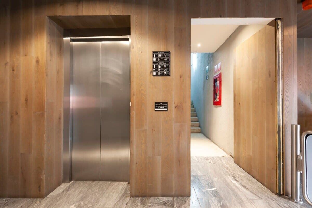 A modern elevator with a metallic finish, flanked by wooden panel walls and a tiled floor.