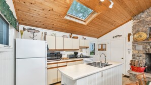 Une cuisine compacte avec des meubles blancs, un îlot central doté d’un évier, une cheminée en pierre et une lucarne.