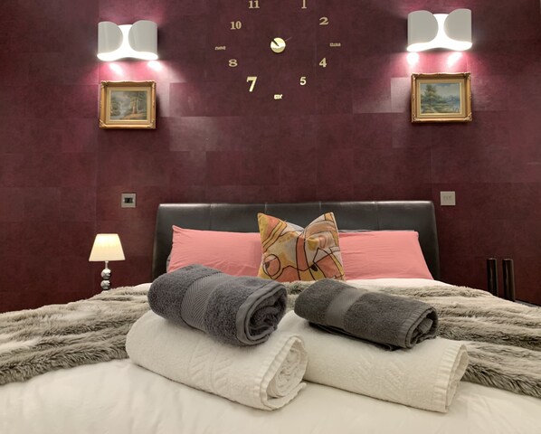 A neatly made bed with two rolled towels, a pink pillow, and a patterned pillow on a dark headboard.