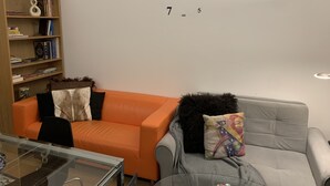 Un salon avec un canapé orange, un canapé gris, une table basse en verre et une bibliothèque adossée au mur.