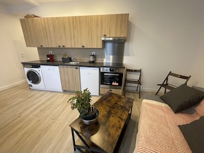 Une cuisine compacte équipée d’un lave-linge, d’un lave-vaisselle et d’un four. On y trouve une table en bois avec une plante en pot et un canapé recouvert d’une couverture à motifs.
