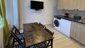 Cocina compacta con lavadora integrada, una mesa de comedor con sillas y un televisor de pantalla plana montado en la pared.
