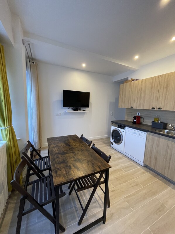Cocina compacta con lavadora integrada, una mesa de comedor con sillas y un televisor de pantalla plana montado en la pared.