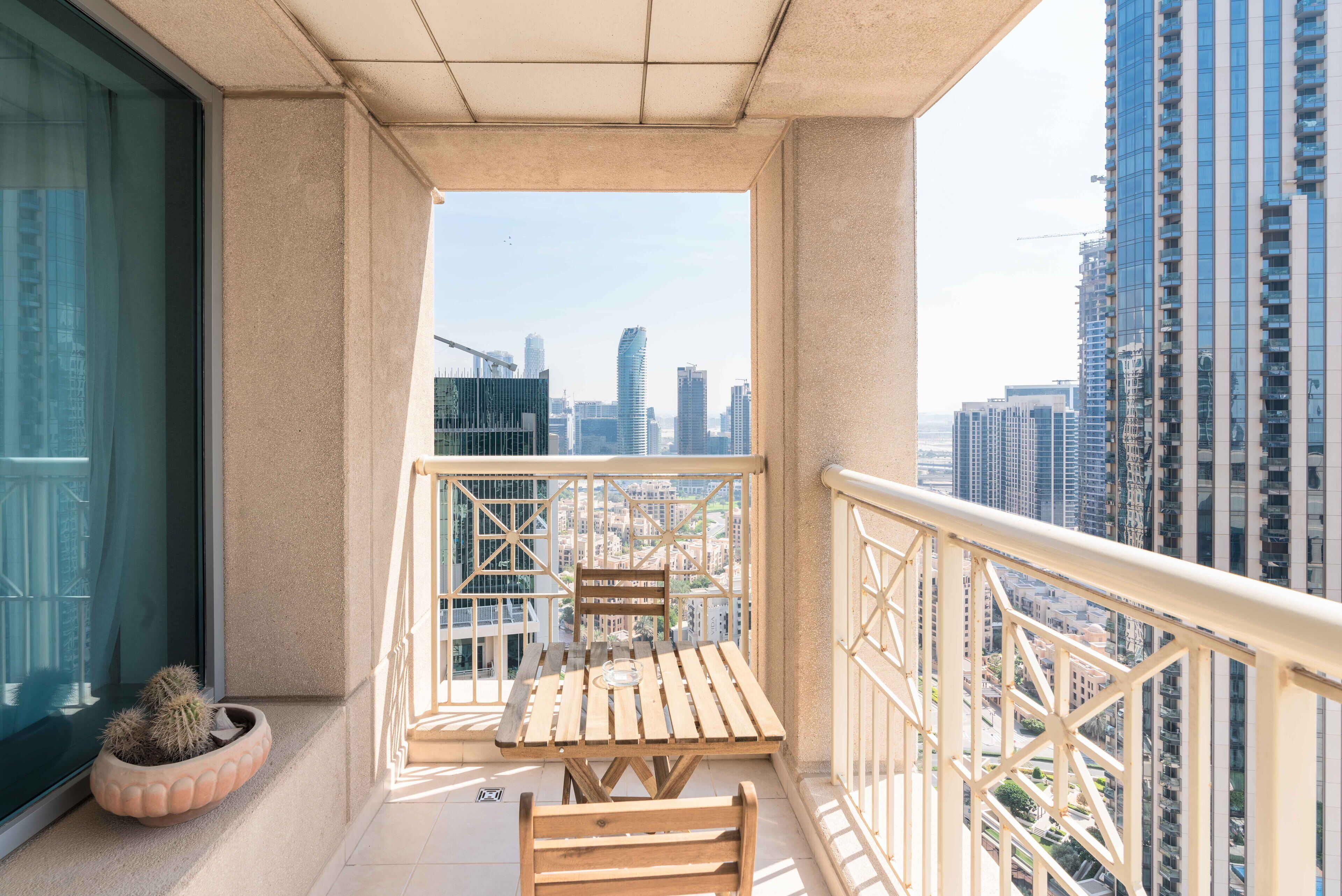Un balcon avec une table et des chaises en bois, un pot de fleurs et une vue sur un paysage urbain dominé par des immeubles de grande hauteur.