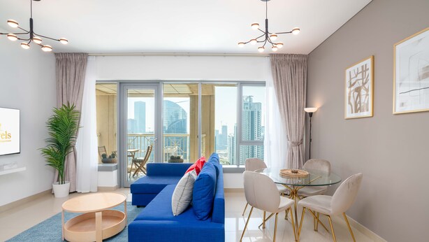 A modern living room with a blue sofa, a glass dining table with white chairs, and a large window with a city view. - Lux BnB I 29 Burj BLVD I Downtown Views (Dubai)