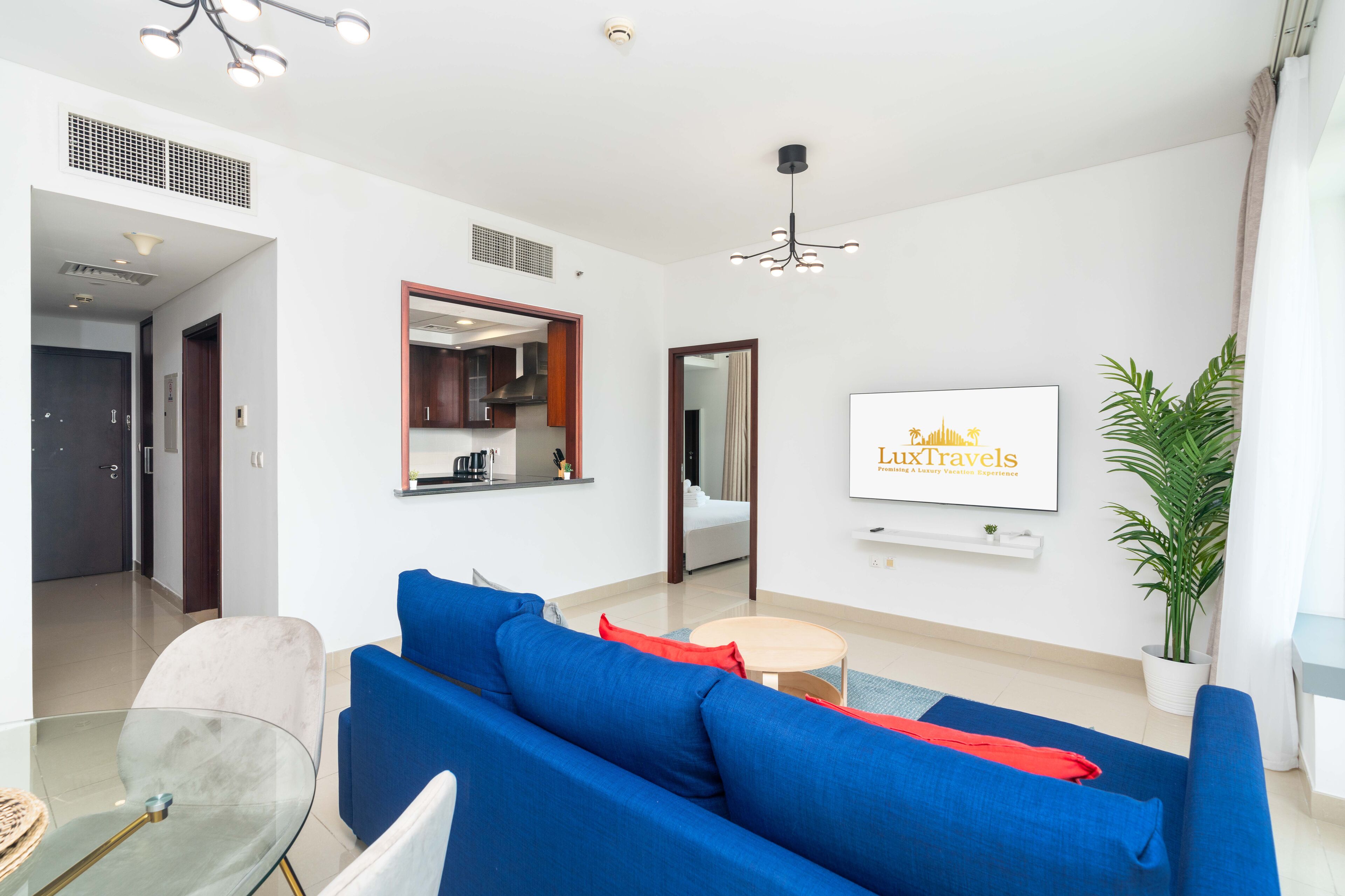 A modern living room with a blue sofa, a glass coffee table, and a dining area with a white table and chairs. A kitchen is visible in the background, and a framed sign on the wall reads "LuxTravels."