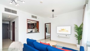 A modern living room with a blue sofa, a glass coffee table, and a dining area with a white table and chairs. A kitchen is visible in the background, and a framed sign on the wall reads "LuxTravels." - Lux BnB I 29 Burj BLVD I Downtown Views (Dubai)