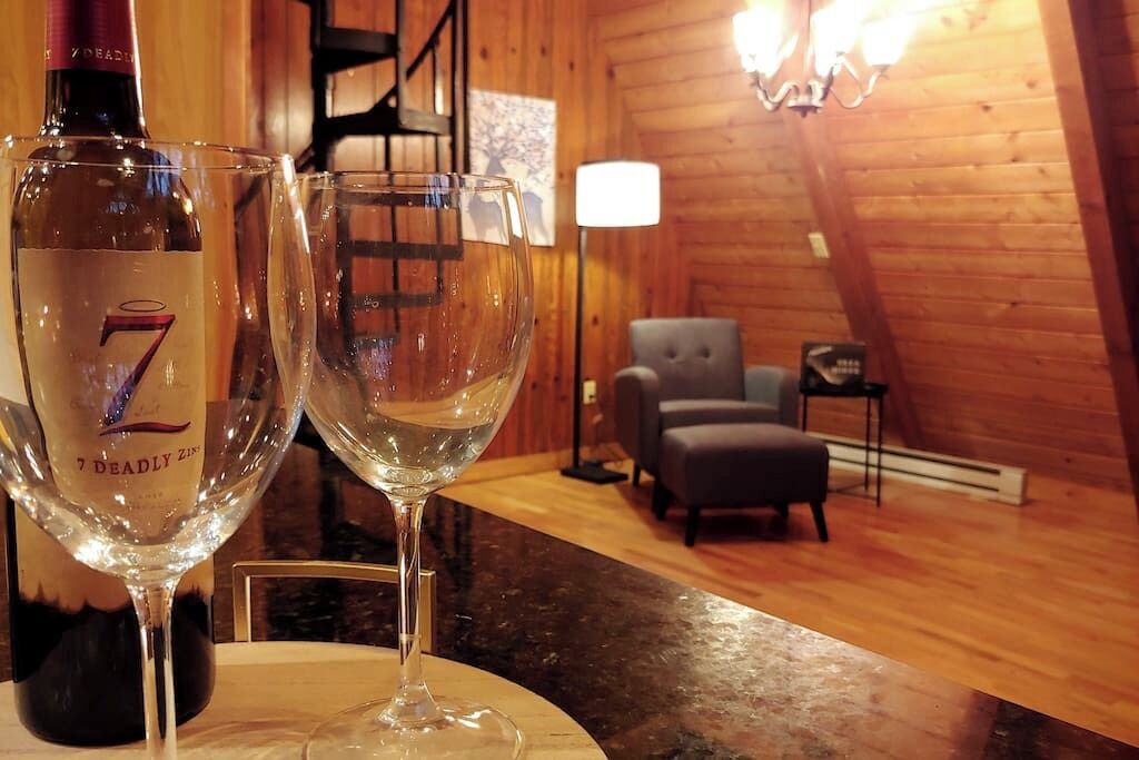 A bottle of 7 DEADLY ZINZICIN and two empty wine glasses on a table in a wooden interior with a chair and a lamp in the background.