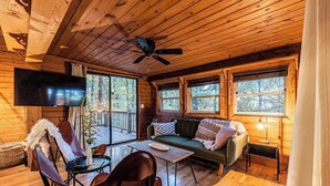 Interior acogedor de una cabaña con techo de madera, ventilador de techo, televisor montado en la pared, sofá verde, sillón de cuero marrón, una mesita y ventana con vista a árboles.