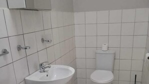 A small, clean bathroom with white tiles, a pedestal sink, a toilet, and a plunger on the floor. - monument pavilion (Gqeberha (Port Elizabeth))