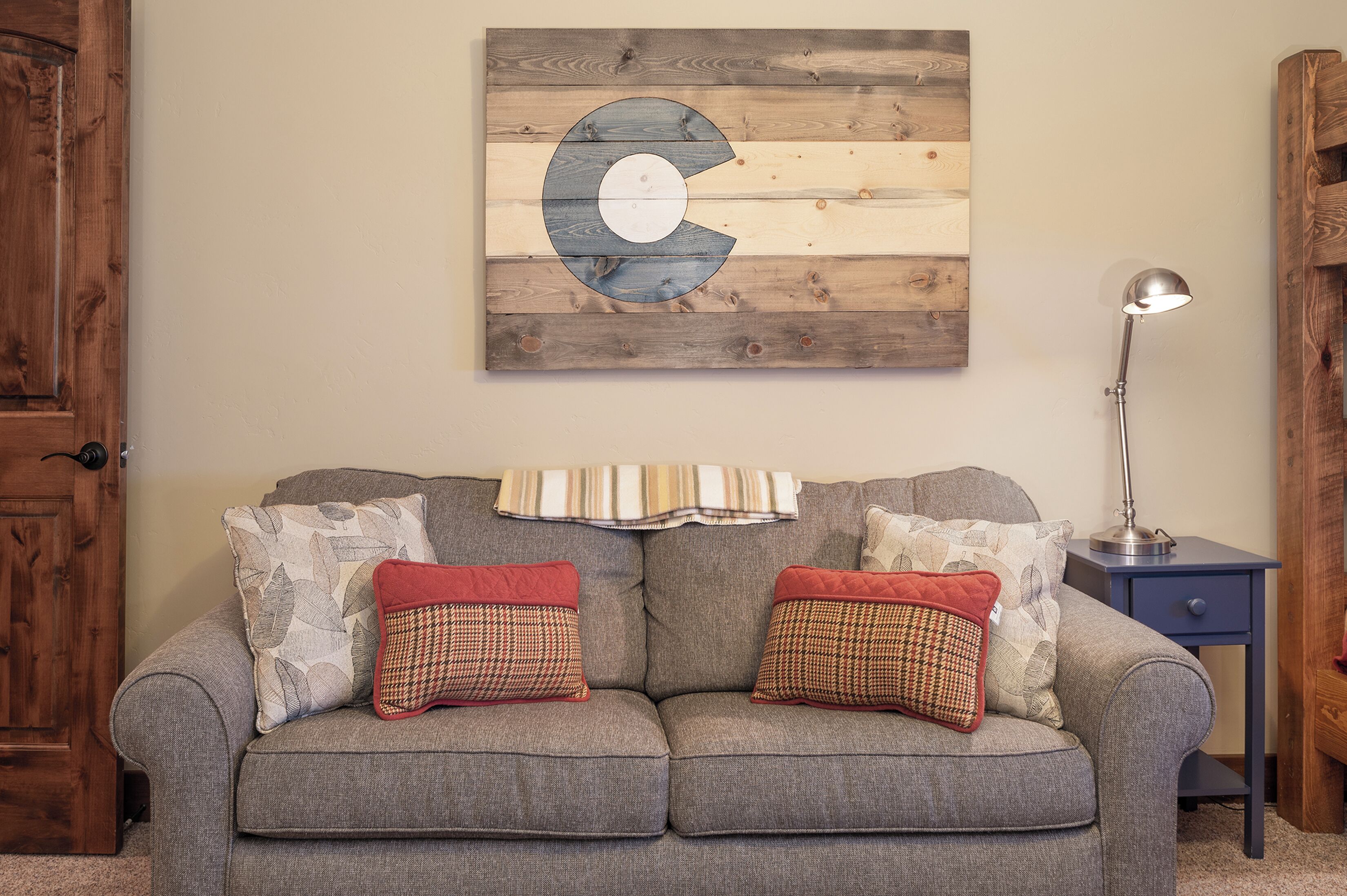 A grey sofa with two patterned cushions, a wooden wall with a large letter C, a striped throw, and a side table with a lamp.