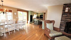 A spacious living area with a dining table, white chairs, a wicker armchair, and a fireplace. - Sprawling Home With Great Outdoor Space (2288) (Harwich Port)