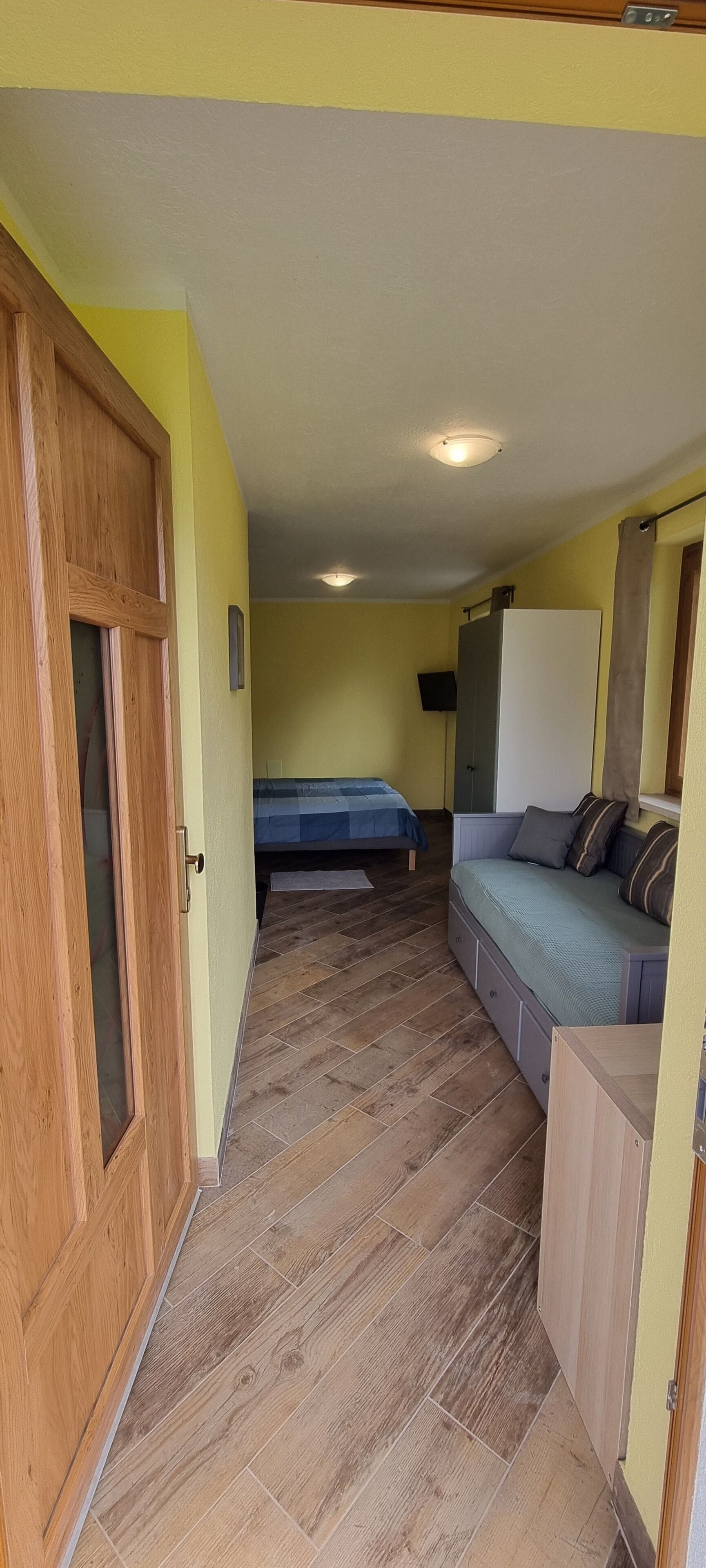 Quarto compacto com porta de madeira, cama com colcha azul, sofá com almofadas e armário branco.