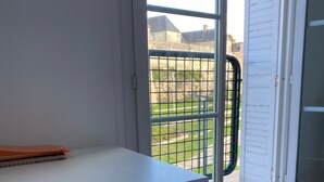 Una stanza con una scrivania e una sedia bianche, una finestra con inferriata metallica e vista su un edificio e del verde all'esterno.
