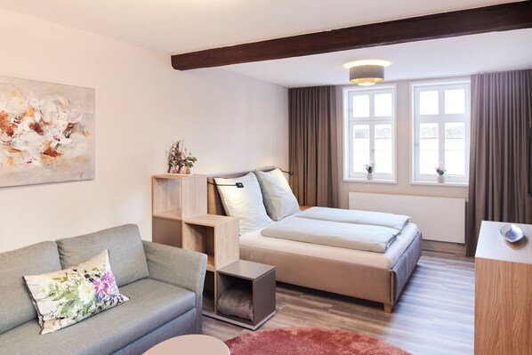Una habitació d'hotel moderna amb un sofà, un llit, una taula auxiliar de fusta i una finestra amb cortines.