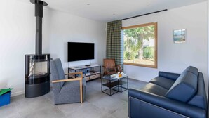 Een moderne woonkamer met een zwart houtgestookte kachel, een flatscreen televisie, een grijze bank en een houten stoel.