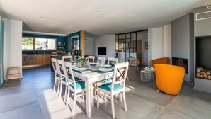 Un comedor moderno con una mesa blanca y sillas azul turquesa, una cocina al fondo y una chimenea con un sillón grande color naranja.