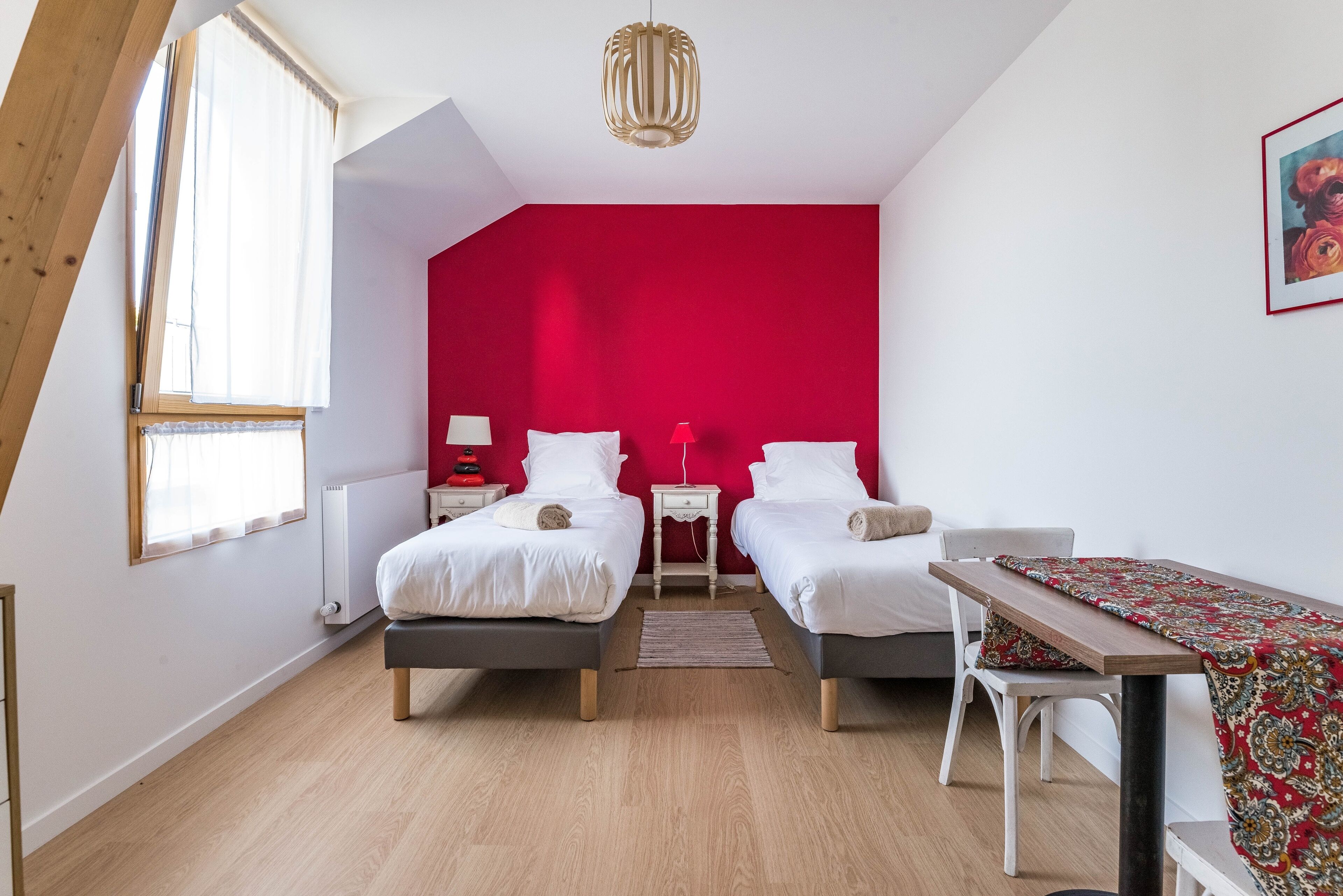 A bedroom with two beds, a red accent wall, a wooden ceiling, a table with a patterned tablecloth, and a window with sheer curtains.