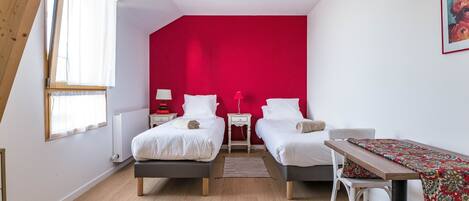A bedroom with two beds, a red accent wall, a wooden ceiling, a table with a patterned tablecloth, and a window with sheer curtains.