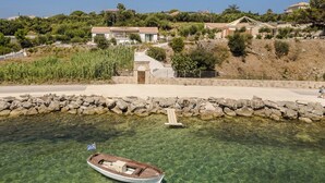 A coastal scene with a clear view of a small boat docked near a rocky shoreline, a stone wall, and houses in the background. - Apollonides Seafront Villa 1 “Aoede” (Kefalonia)