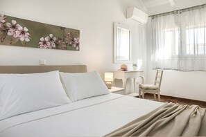 Een netjes opgemaakt bed met witte lakens, een nachtkastje met een lamp, een stoel, een bureau met een plant en een groot bloemschilderij aan de muur.
