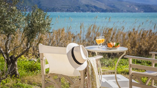 A table with a hat and two glasses of orange juice, overlooking a lake with mountains in the background. - Apollonides Seafront Villa 1 “Aoede” (Kefalonia)