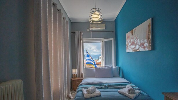 A bedroom with a bed, a window with curtains, a radiator, a bedside table, and a painting on the wall. - Sea View Nikos Apartment (Ναύπλιο)