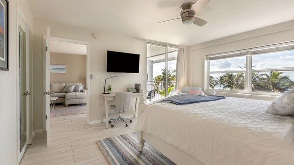 Een slaapkamer met een bed, een bureau, een stoel, een televisie en een balkon met uitzicht op palmbomen.