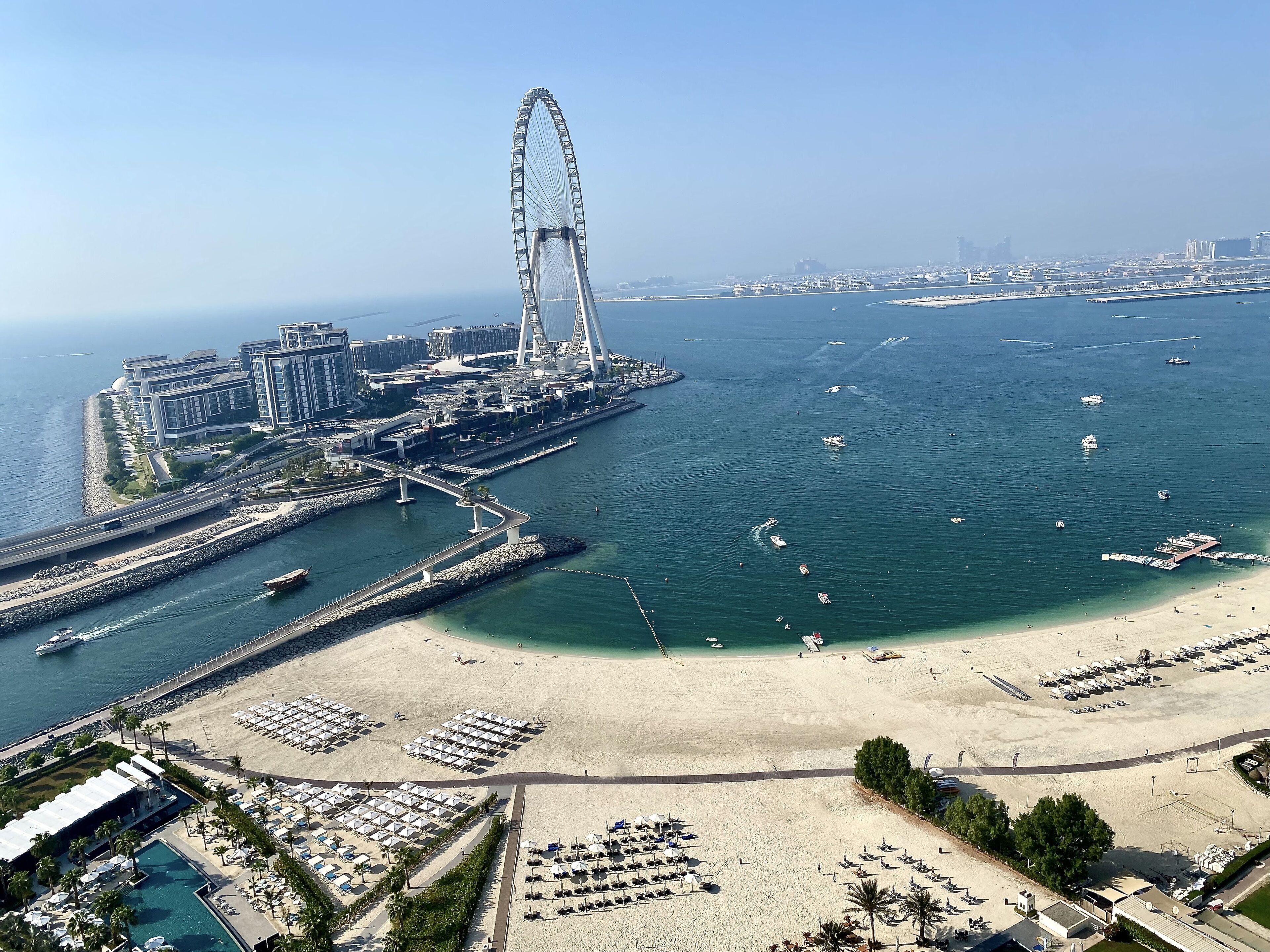 Eine Küstenstadt mit einem markanten Riesenrad, einem Strand mit Liegestühlen und einem Jachthafen mit Booten.