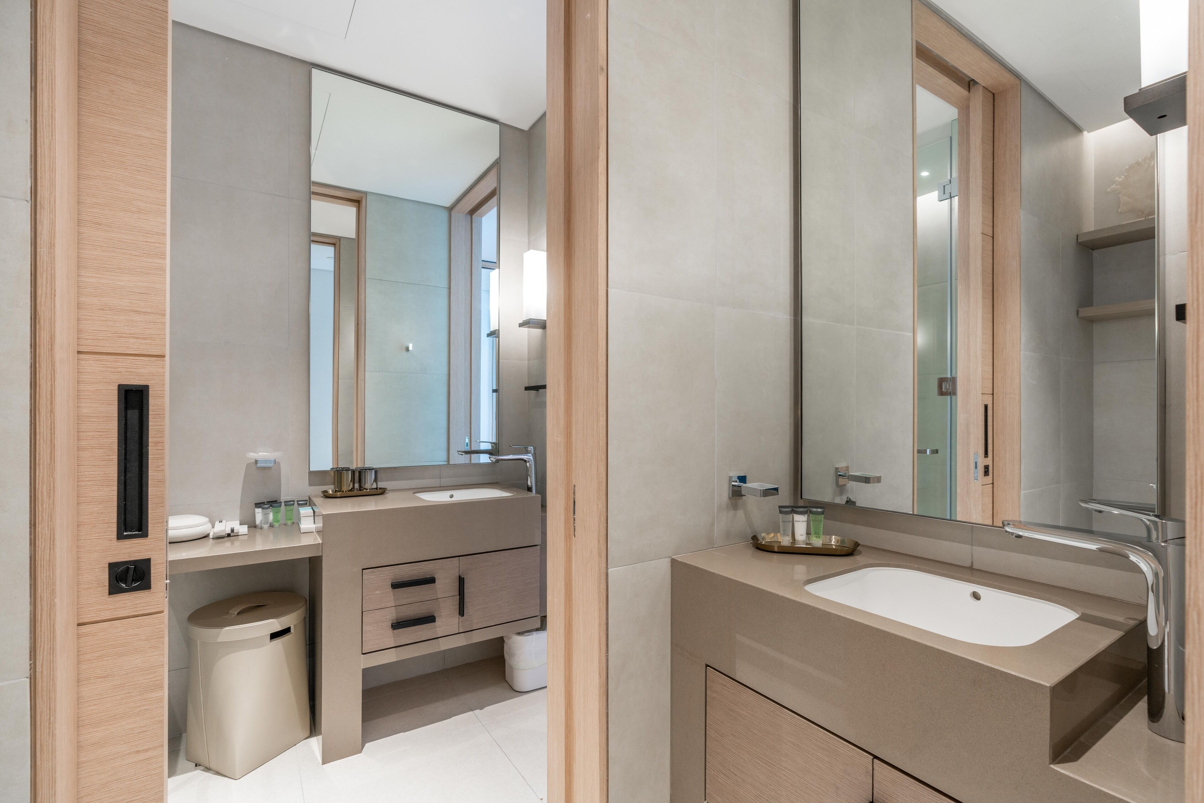A modern bathroom with a large mirror, two sinks, a vanity, and a trash bin.