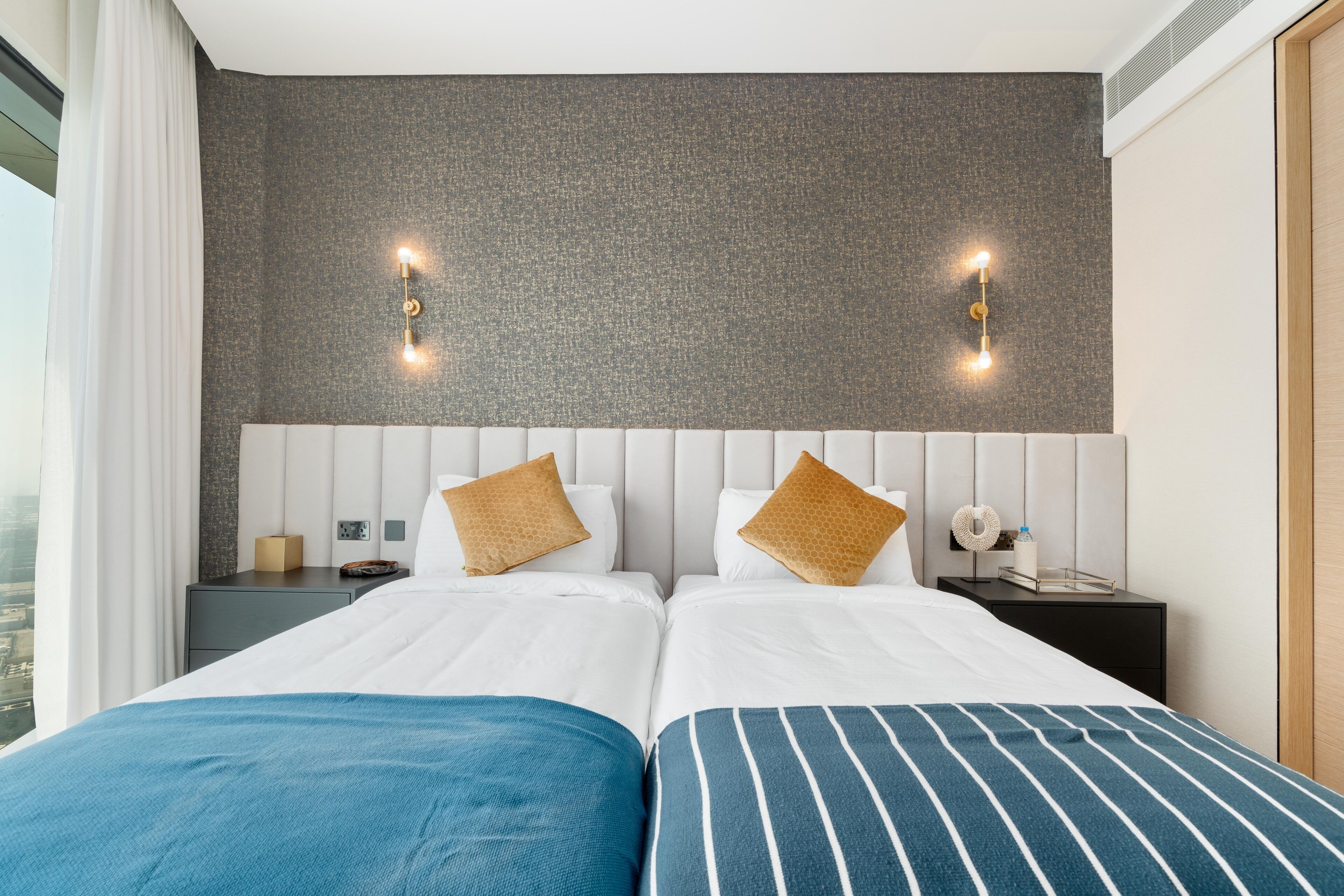 Une chambre d’hôtel avec deux lits, un mur gris à texture et une vue sur la ville.