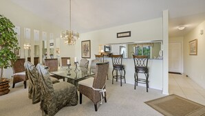 A dining area with a glass table surrounded by wicker chairs, a chandelier, and a potted plant. - Condo in Golf Community (Naples)