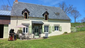 A two-story house with a sloped roof, green shutters, and a small patio area with outdoor furniture. - 1802 Auvergne house (Saint-Donat)