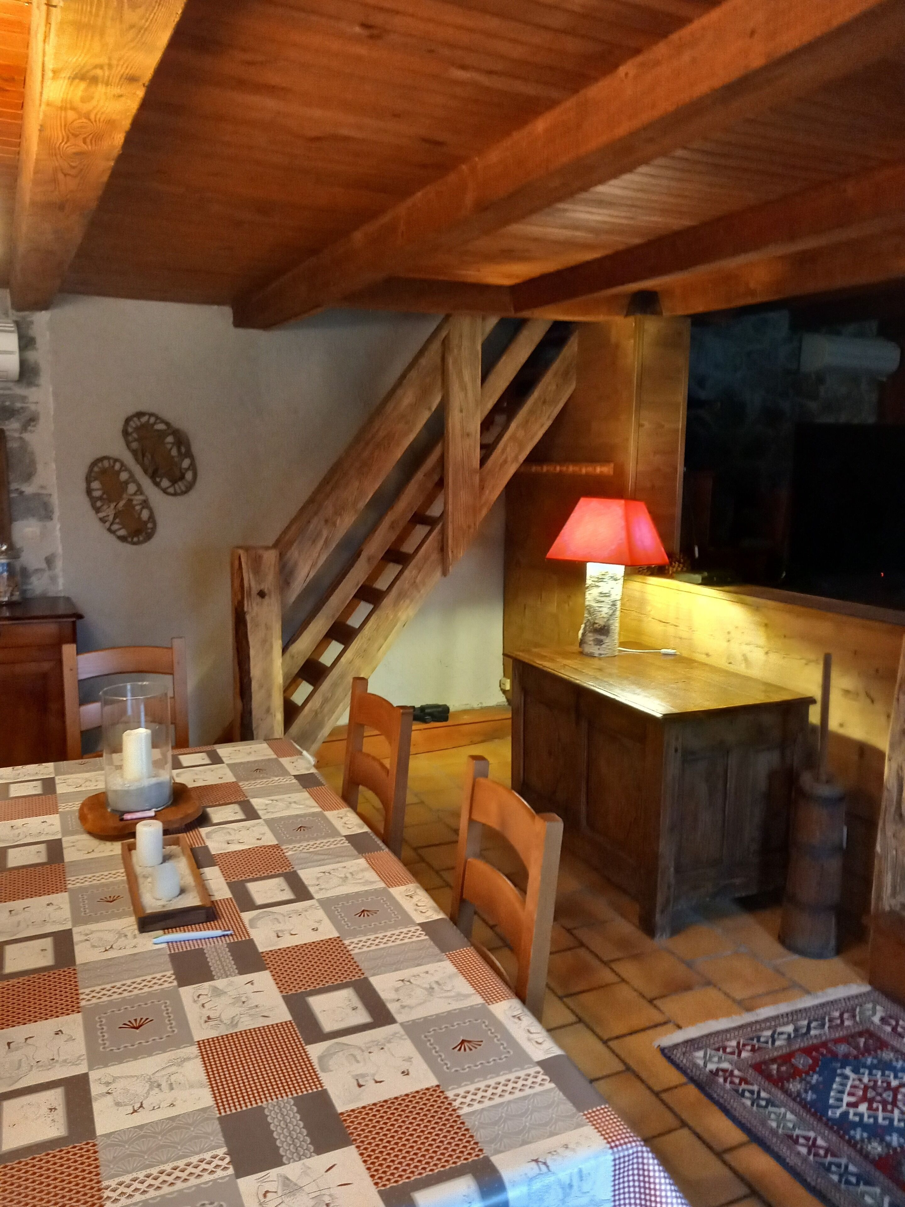 Un'accogliente zona pranzo con un tavolo in legno, sedie e una tovaglia trapuntata. Una scala conduce a un livello superiore, e una lampada rossa è visibile sulla parete.