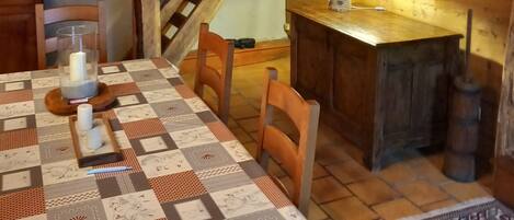 Un coin repas accueillant avec une table en bois, des chaises et une nappe matelassée. Un escalier mène à un niveau supérieur, et une lampe rouge est visible sur le mur.