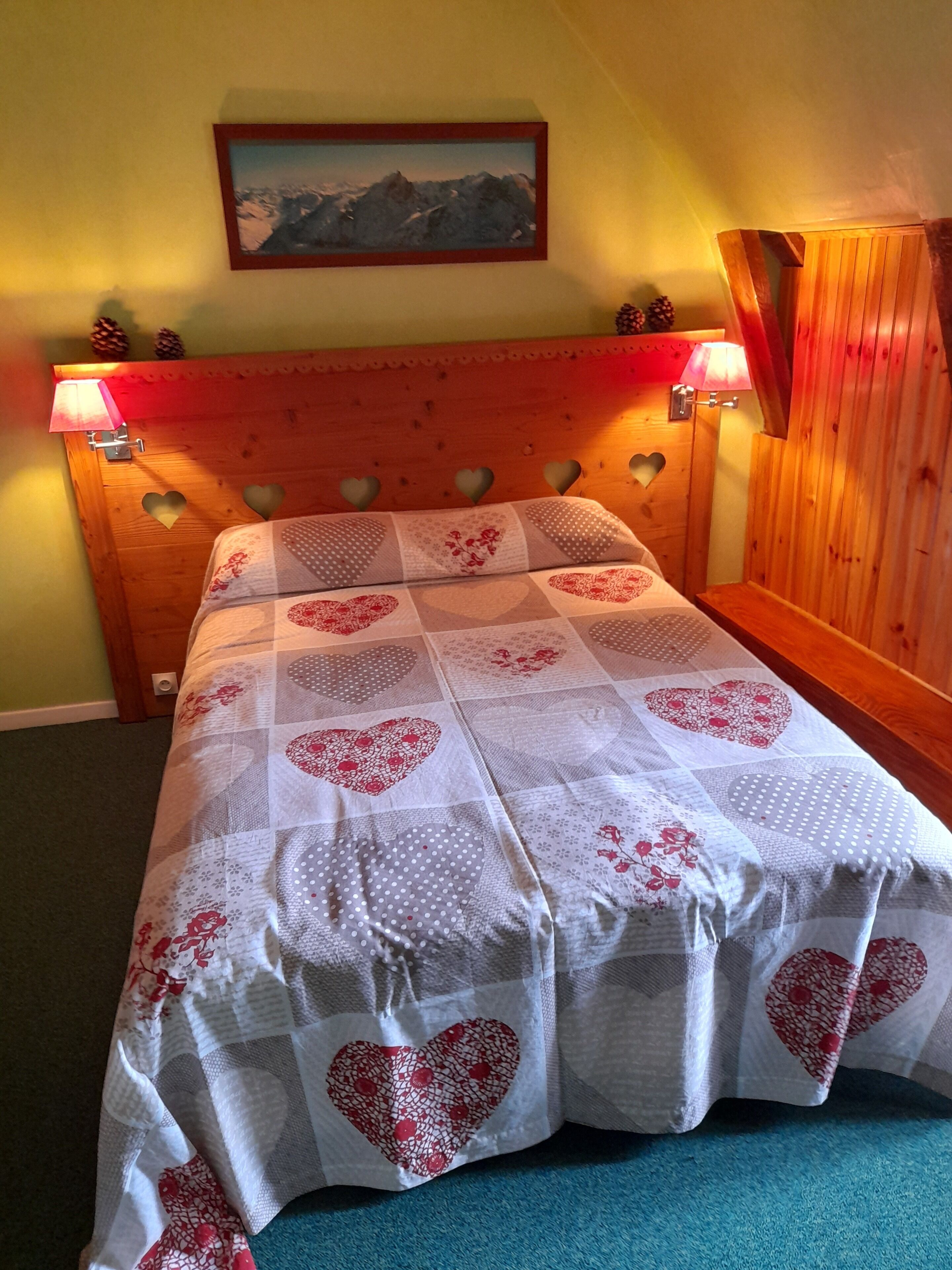 Une chambre à coucher avec une tête de lit en bois, une literie à motifs de cœurs et un tableau représentant un paysage de montagne au-dessus du lit.