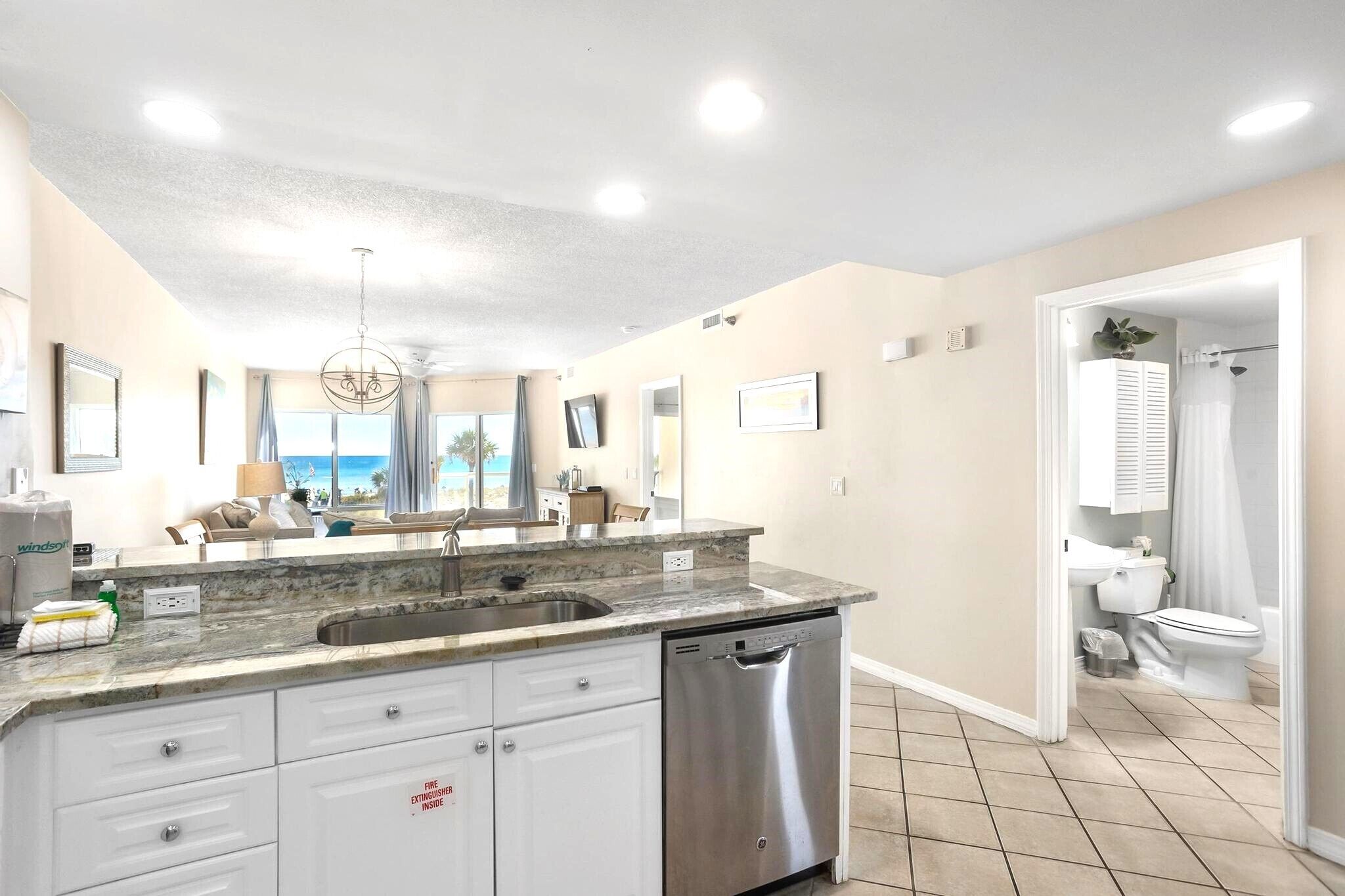 A modern kitchen with a marble countertop, stainless steel dishwasher, and a view of the ocean through large windows.