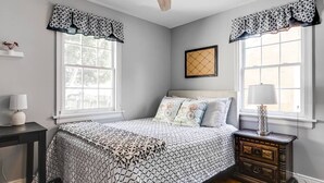 Un dormitorio con cama, mesitas de noche, ventilador de techo y ventanas con cortinas decoradas.