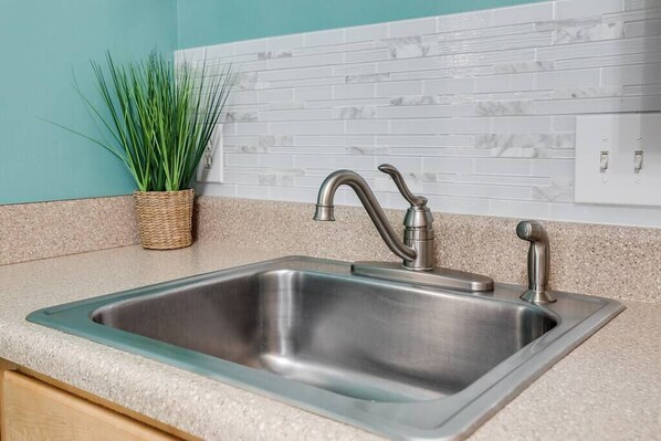 A kitchen with a stainless steel sink, a woven basket with green plants, and a white tiled backsplash.