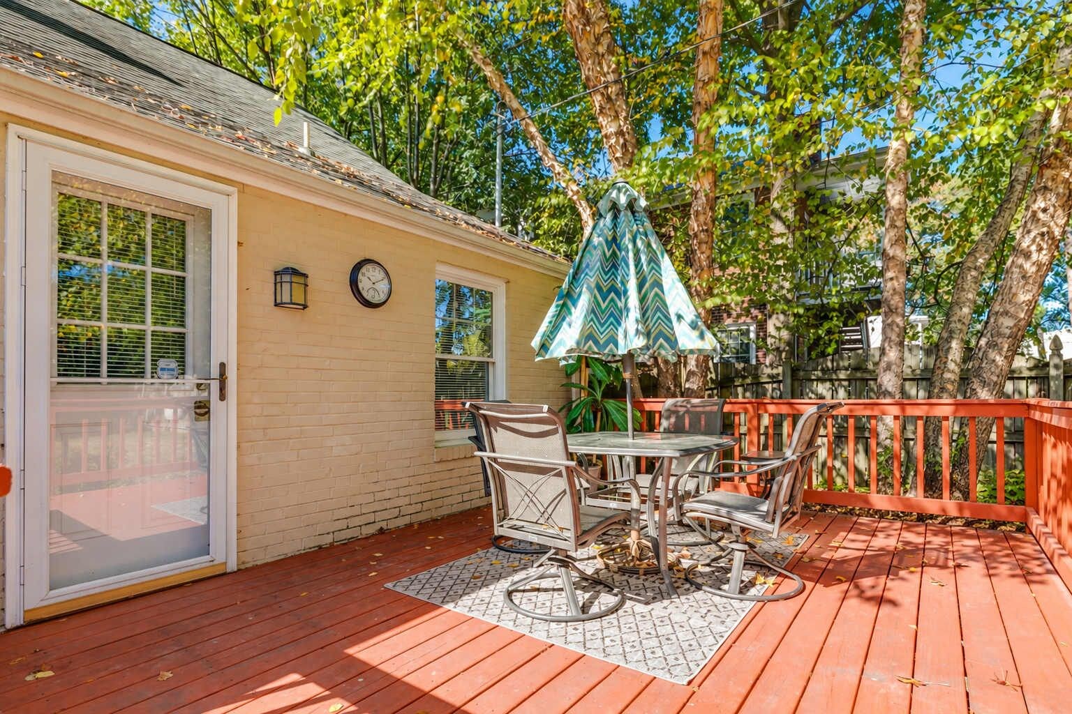 Une terrasse avec une table et des chaises, une horloge au mur et une rambarde rouge.
