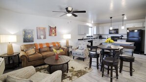 A modern living room with a sofa, coffee table, and chairs, a kitchen with white cabinets and appliances, and a ceiling fan. - CK Stay Hotel (Butte)