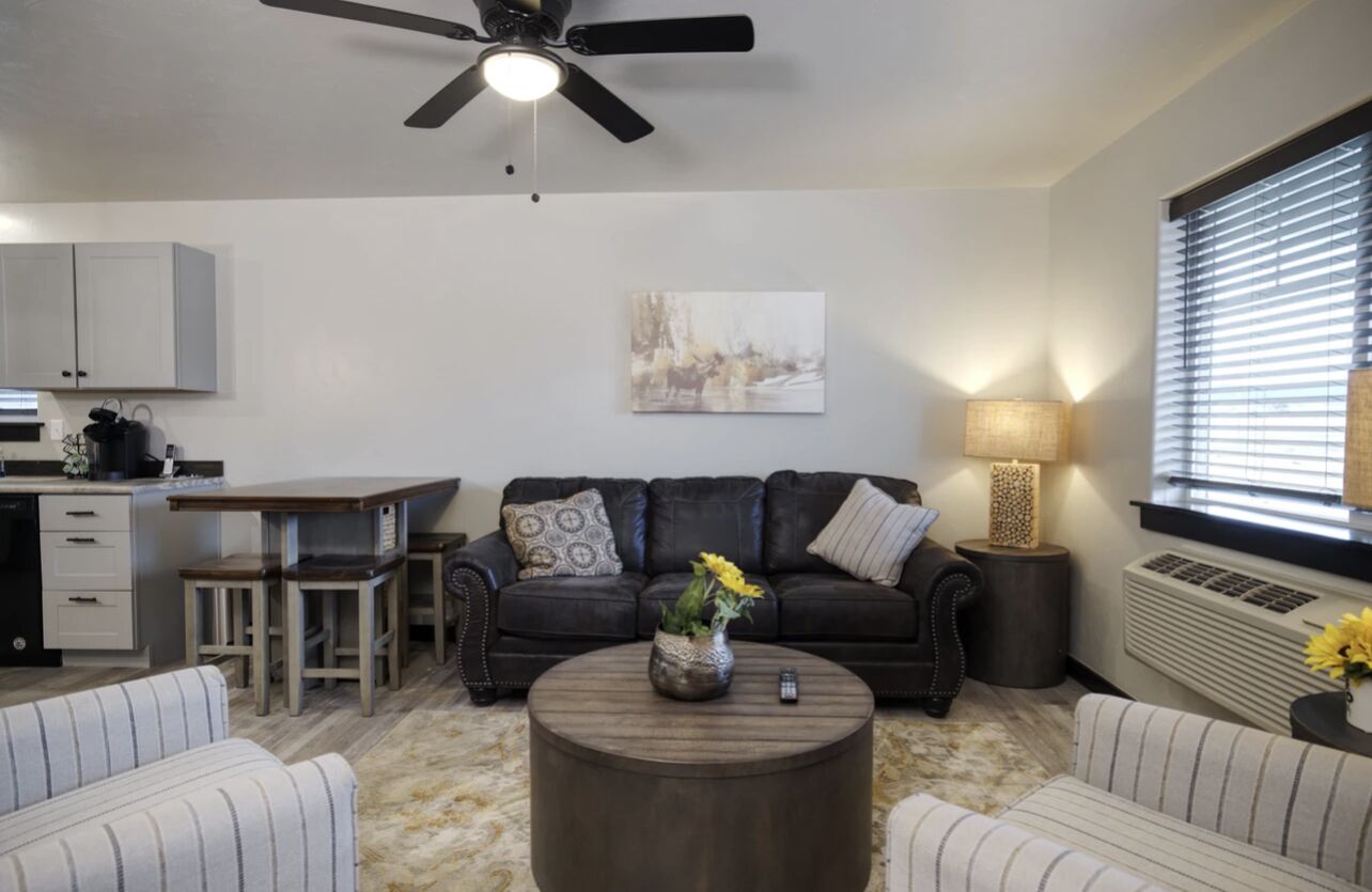 A living room with a sofa, coffee table, and a ceiling fan. There is a dining table with chairs and a kitchen area with cabinets.