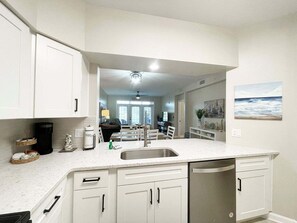 A modern kitchen with white cabinetry, a stainless steel sink, and a dishwasher. There is a dining area in the background with a view of the ocean. - Seaside at Anastasia B102 (St. Augustine)