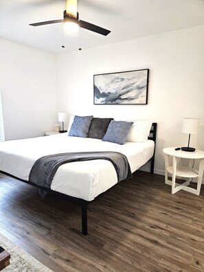 A bedroom with a bed, a ceiling fan, a bedside table, a nightstand, and a wall-mounted artwork.