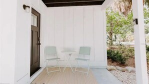 Une terrasse couverte avec deux chaises et une table, une porte sombre et un mur extérieur blanc.