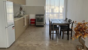 Eine kleine Küche mit Kühlschrank, Herd und Spüle. Ein Esstisch mit Stühlen und ein Blumentopf sind ebenfalls vorhanden.