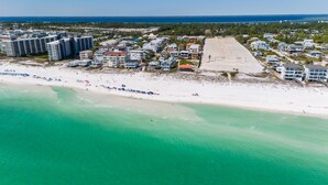 Vue aérienne d’une zone côtière avec une plage de sable, un grand parking et plusieurs immeubles à étages.