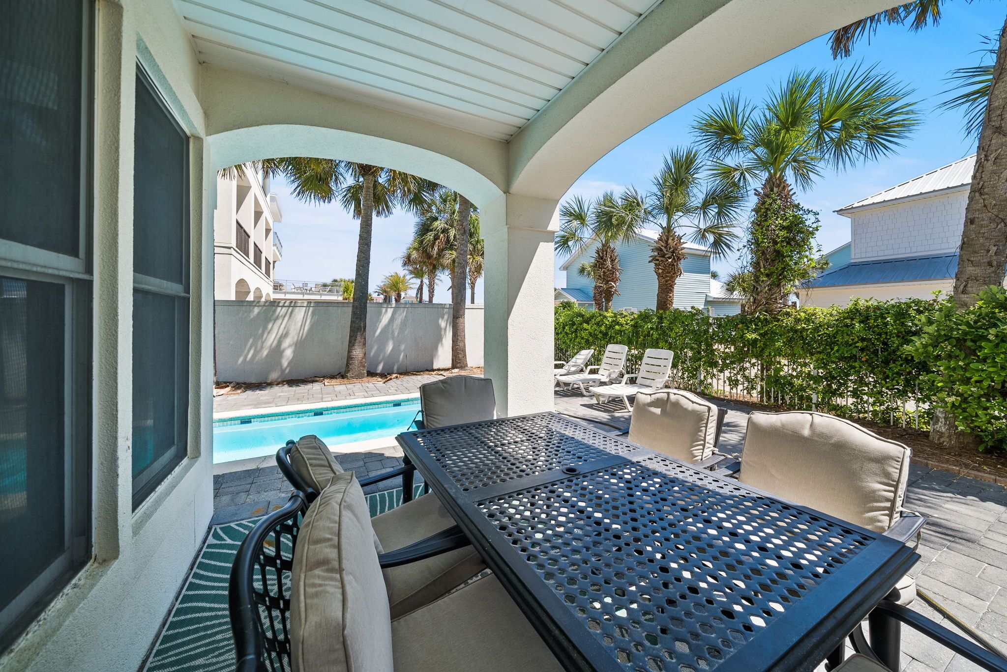 A covered outdoor seating area with a pool, lounge chairs, and palm trees.
