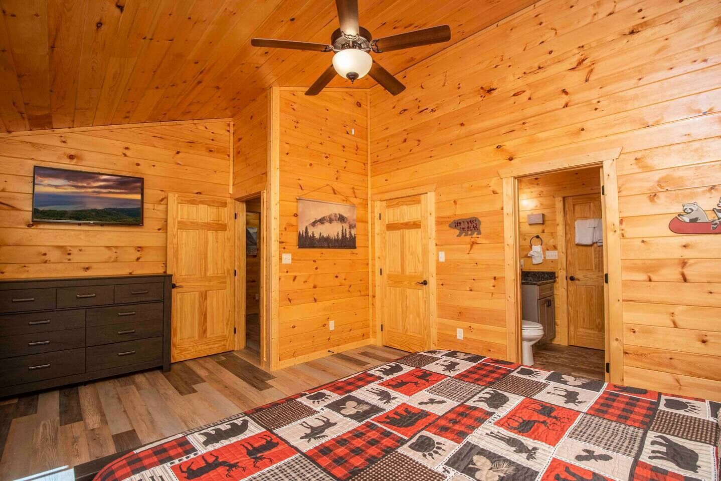 A wooden cabin interior with a bed, a dresser, a ceiling fan, and a painting on the wall.