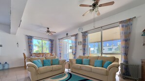 Ein geräumiges Wohnzimmer mit zwei beigefarbenen Sofas, einem Teppich mit blau-schwarzem Muster und Deckenventilatoren.